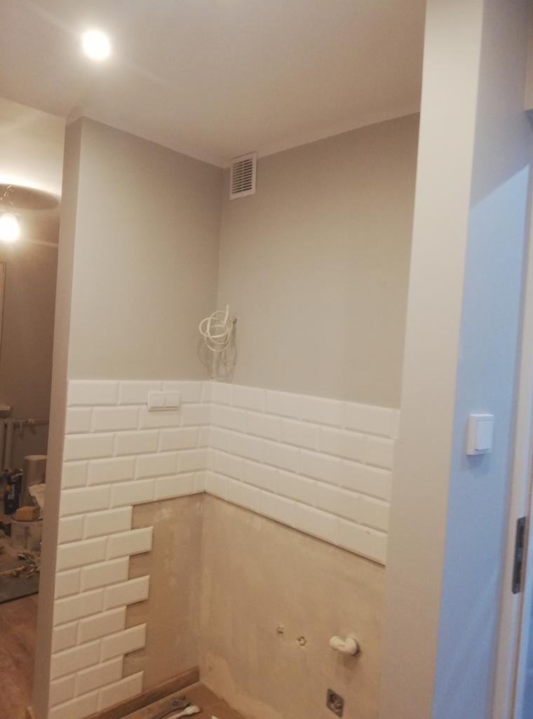 Ściana łazienki w trakcie wykańczania, widoczne białe, prostokątne płytki ułożone do połowy wysokości, odsłonięty fragment ściany z widocznymi instalacjami elektrycznymi oraz fragment podłogi...