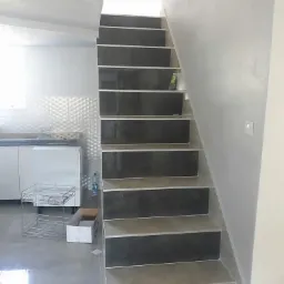 Wykończone schody wewnętrzne z ciemnymi płytkami na stopniach i jasnymi podstopnicami, widoczne elementy kuchni w tle, wnętrze w trakcie wykańczania.