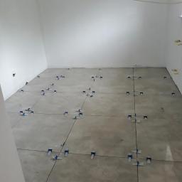 Krzysztof Kuchta - Surowy betonowy podkład w pustym pokoju z białymi ścianami, przygotowany do układania płytek z użyciem systemu poziomowania płytek z niebieskimi klinami i czarnymi krzyżykami dystansowymi.