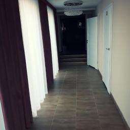 Krzysztof Kuchta - Korytarz z brązowymi płytkami podłogowymi, białymi drzwiami i zasłonami w odcieniach bieli i bordo, oświetlony dwoma kryształowymi żyrandolami.