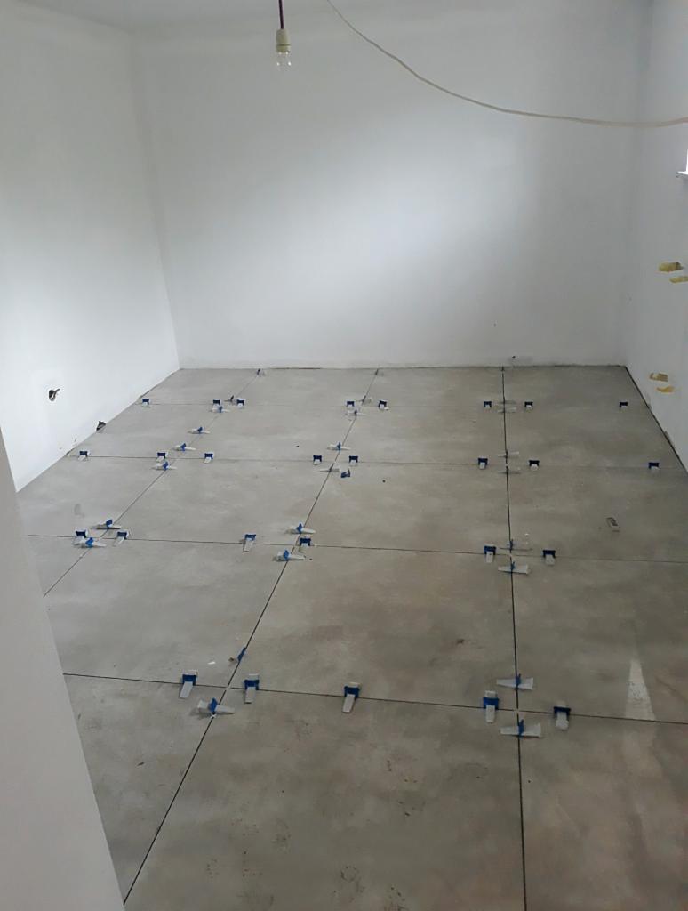 Surowy betonowy podkład w pustym pokoju z białymi ścianami, przygotowany do układania płytek z użyciem systemu poziomowania płytek z niebieskimi klinami i czarnymi krzyżykami dystansowymi.