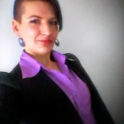 Uśmiechnięta kobieta w czarnej marynarce i fioletowej koszuli, z krótkimi włosami i kolczykami w kształcie kół, pozuje na jasnym tle.