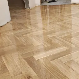 Świeżo polakierowany parkiet ułożony we wzór jodełki, widoczne odbicia światła na mokrej powierzchni, pomieszczenie w trakcie remontu z widocznym bałaganem w tle.