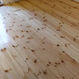 Fragment drewnianej podłogi z widoczną różnicą w kolorze po częściowym usunięciu warstwy lakieru, grzejnik na ścianie w tle.