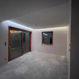 Surowe wnętrze z gładzią, oświetleniem LED w suficie podwieszanym, brązowymi ramami okiennymi i betonową posadzką z plamami. Widoczne niedokończone instalacje elektryczne.