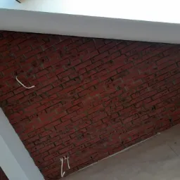 Ściana wykończona czerwoną cegłą klinkierową z widocznymi przewodami elektrycznymi wystającymi ze ściany, surowy beton na podłodze.