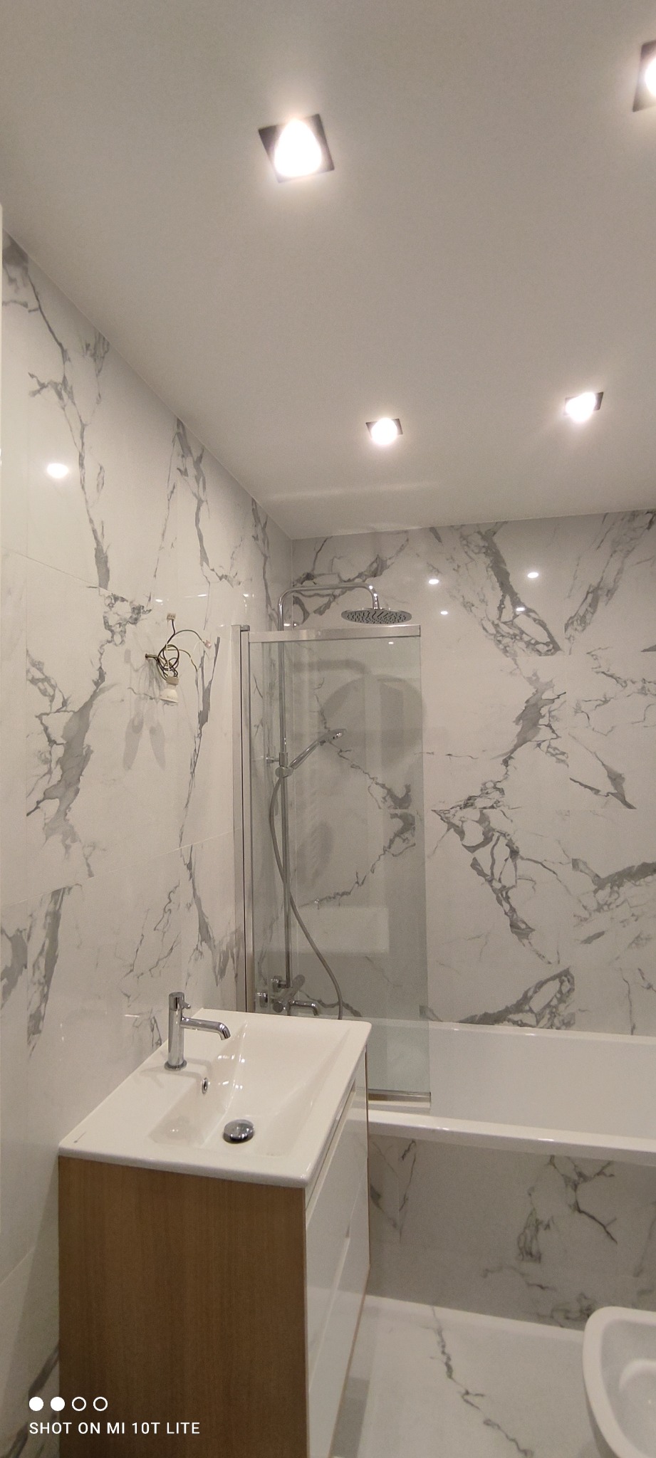 Nowoczesna łazienka z marmurowymi płytkami, kabiną prysznicową za szklaną ścianką i umywalką z szafką w kolorze drewna.