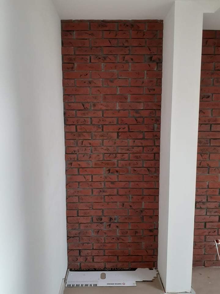 Czerwona ściana z cegły dekoracyjnej między białymi ścianami, widoczna kartonowa podkładka pod panele na podłodze.