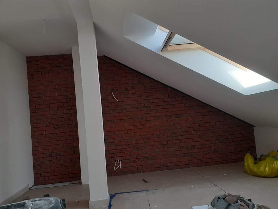 Poddasze w trakcie prac wykończeniowych: ściana wyłożona czerwoną cegłą, biała kolumna i okno dachowe w skosie, widoczne materiały budowlane na podłodze.