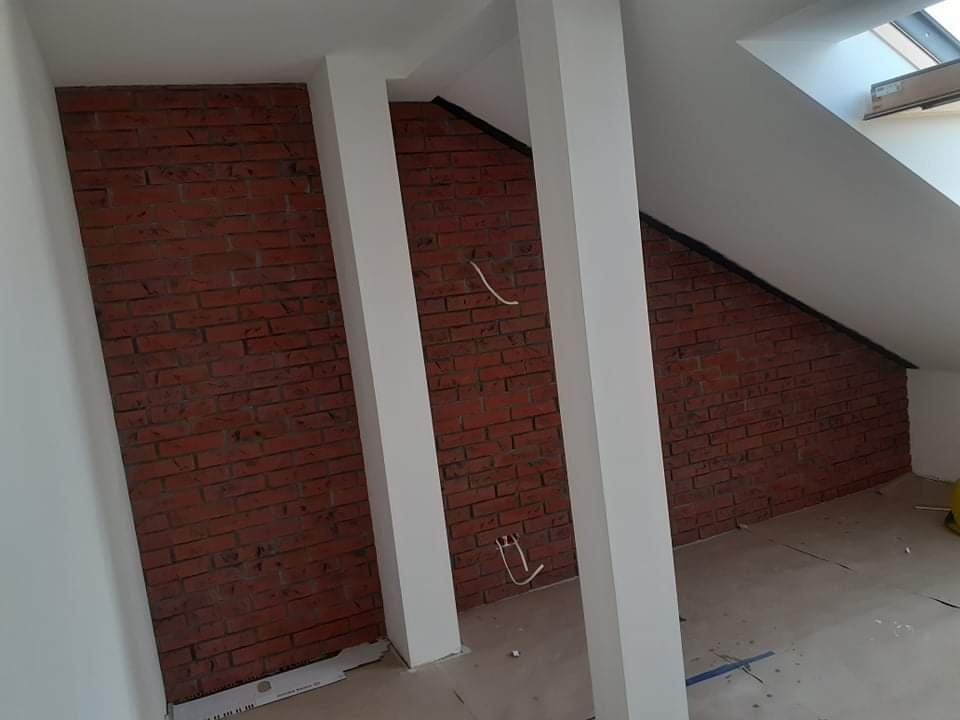 Poddasze po remoncie z fragmentami ściany wykończonej czerwoną cegłą, białe filary i ściany, widoczne kable elektryczne i okno dachowe.