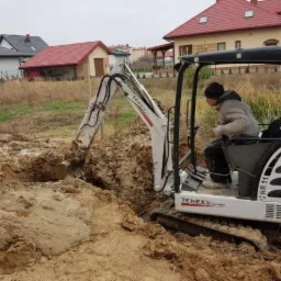 Operator w jasnej kurtce obsługuje białą mini-koparkę Terex HR11 podczas wykopu w grząskim, błotnistym gruncie na tle domów jednorodzinnych.