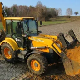 Żółta koparko-ładowarka John O'Flynn B60 z pełną łyżką piasku na polu uprawnym w słoneczny dzień.