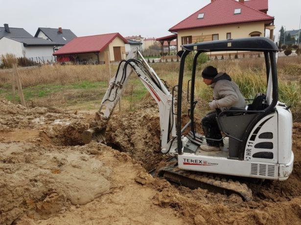 Operator w jasnej kurtce obsługuje białą mini-koparkę Terex HR11 podczas wykopu w grząskim, błotnistym gruncie na tle domów jednorodzinnych.