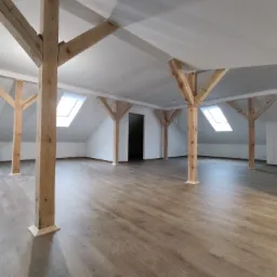 Jasne poddasze z drewnianymi słupami konstrukcyjnymi, nową podłogą z paneli i oknami dachowymi, widok na puste wnętrze.