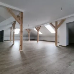 Poddasze po remoncie z drewnianymi belkami, panelami podłogowymi w odcieniach szarości i oknami dachowymi wpuszczającymi jasne światło, widoczne niedokończone punkty oświetleniowe.
