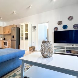 FORM.A MACIEJ APTOWICZ - Jasne wnętrze salonu z aneksem kuchennym, niebieska sofa, biały stolik kawowy z dekoracyjnym lampionem, telewizor na ścianie z dekoracyjnymi talerzami, widoczna zabudowa kuchenna i szafka...