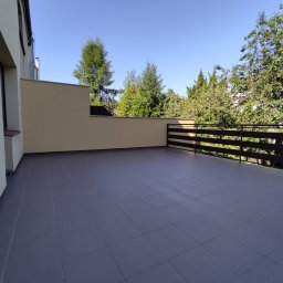 Maciej Aptowicz Usługi Ogólnobudowlane - Przestronny balkon z szarymi płytkami, metalową balustradą i widokiem na zieleń drzew w słoneczny dzień.