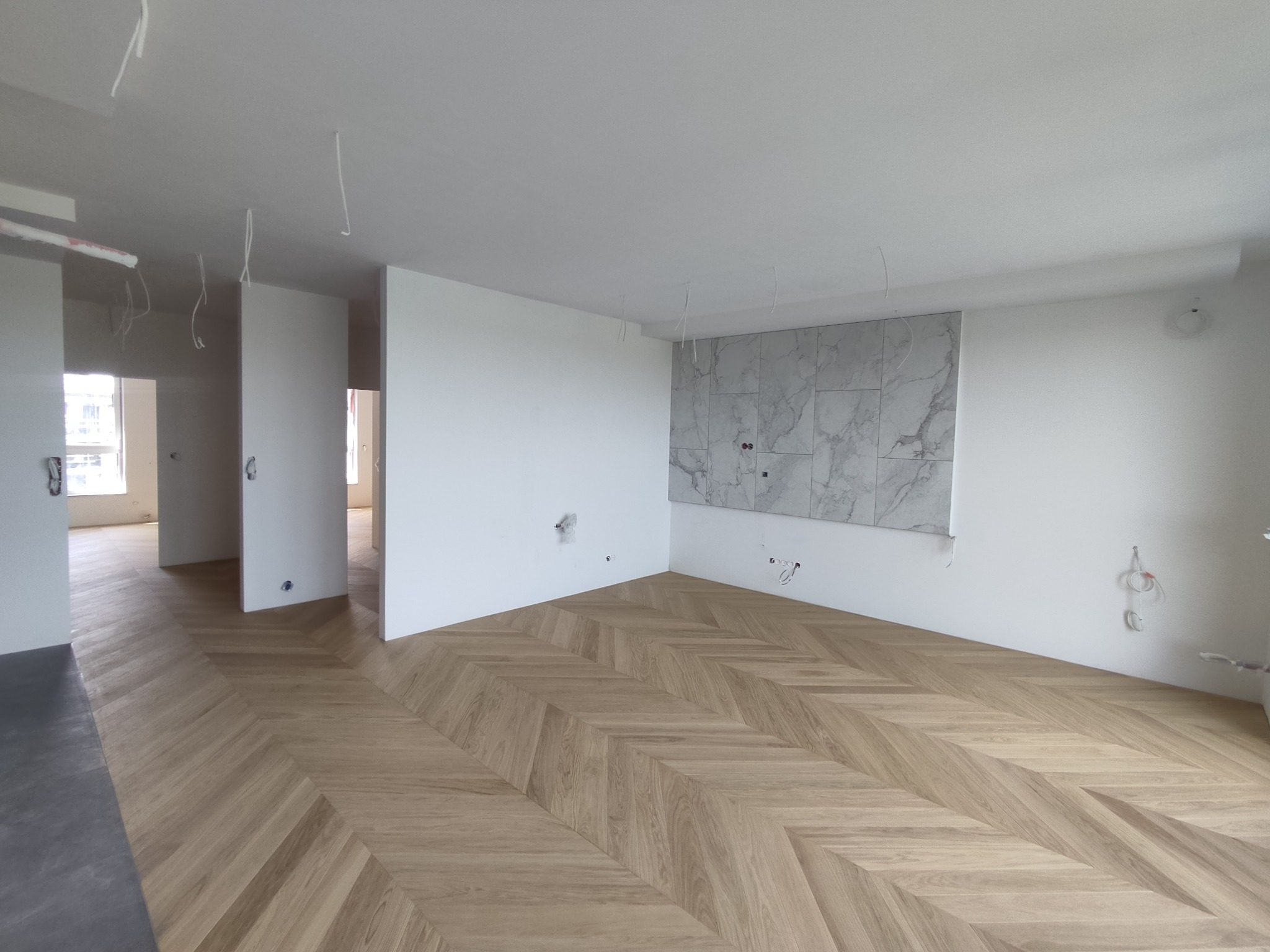 Jasne, przestronne wnętrze w Poznaniu z ułożoną parkietową podłogą w jodełkę, białe ściany z fragmentem wykończonym marmurową płytką, widoczne kable wystające ze ściany i sufitu.