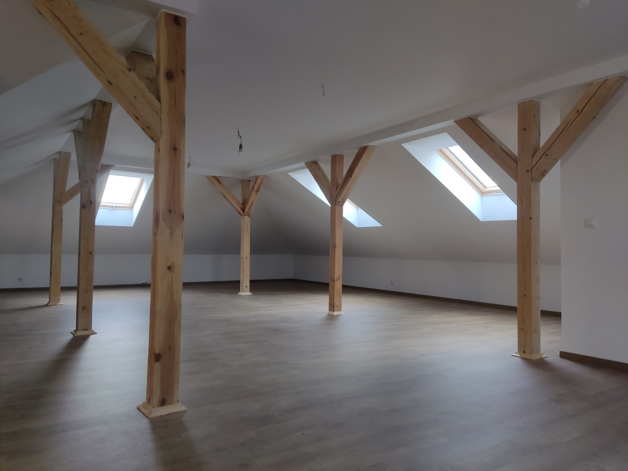 Poddasze po remoncie z widocznymi drewnianymi słupami konstrukcyjnymi, panelami podłogowymi w odcieniach brązu i oknami dachowymi w skosach.