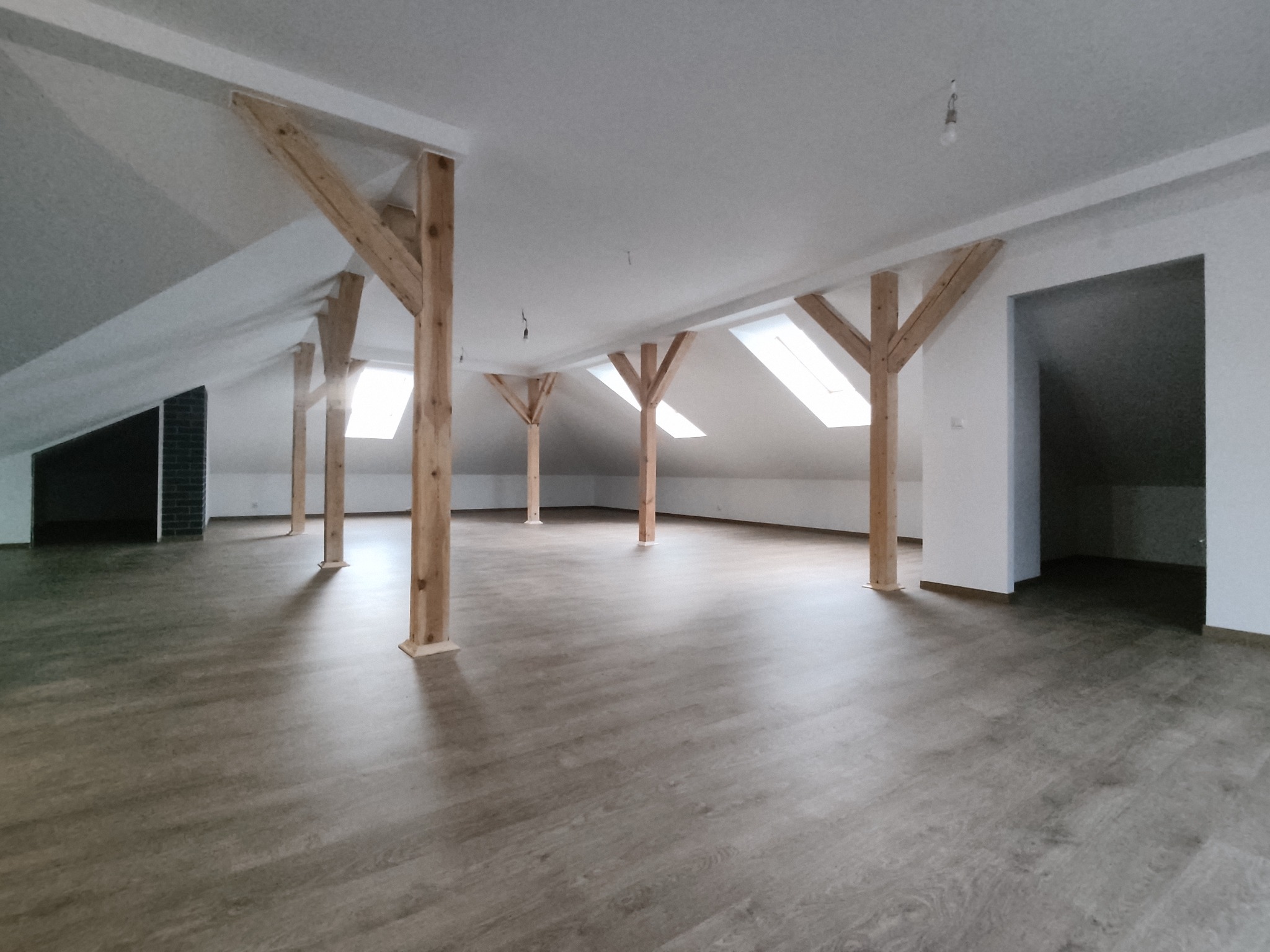 Poddasze po remoncie z drewnianymi belkami, panelami podłogowymi w odcieniach szarości i oknami dachowymi wpuszczającymi jasne światło, widoczne niedokończone punkty oświetleniowe.