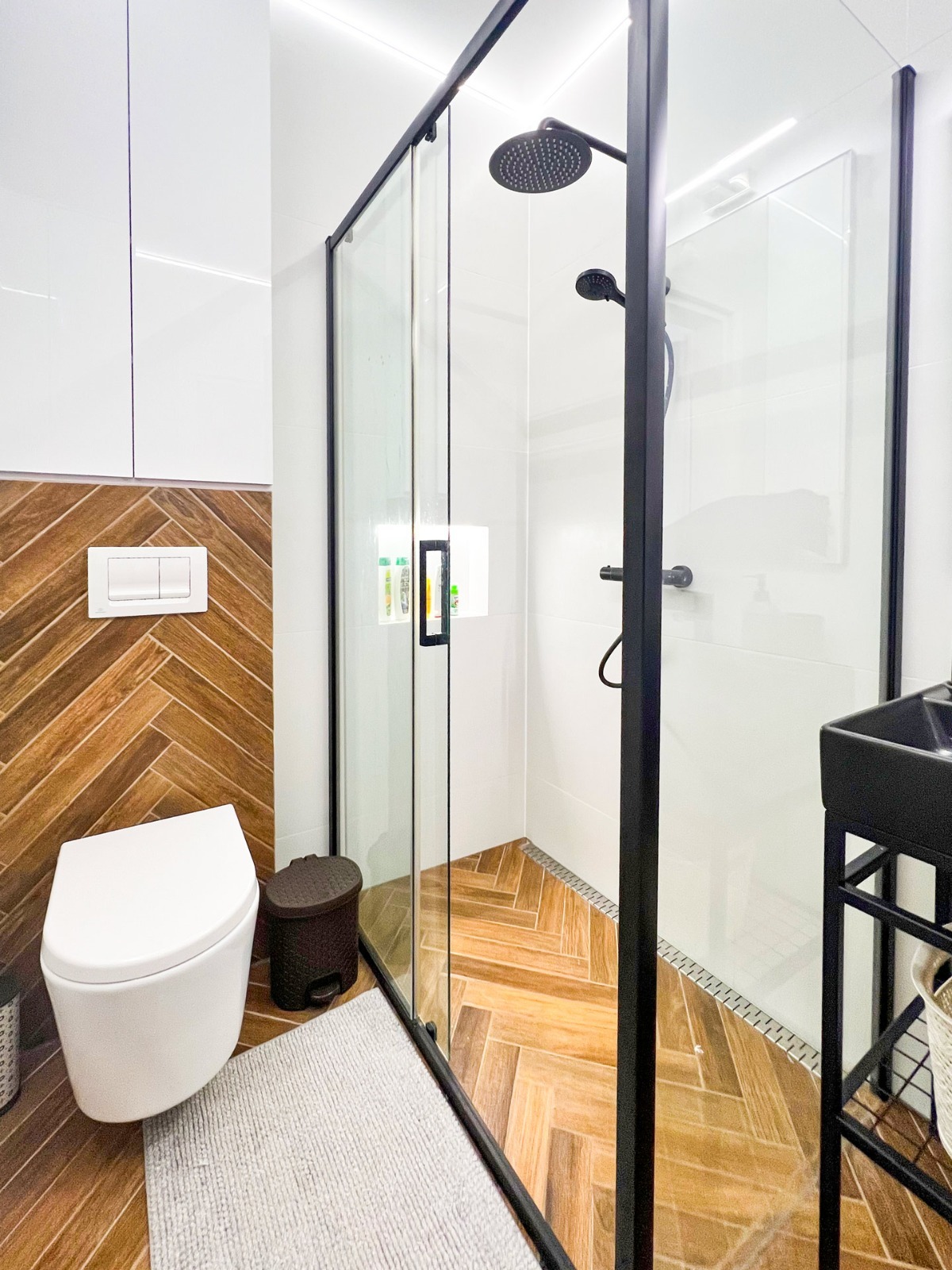 Nowoczesna łazienka z kabiną prysznicową typu walk-in z czarnymi ramami, toaletą wiszącą i brązowymi płytkami ułożonymi w jodełkę na podłodze i częściowo na ścianie.