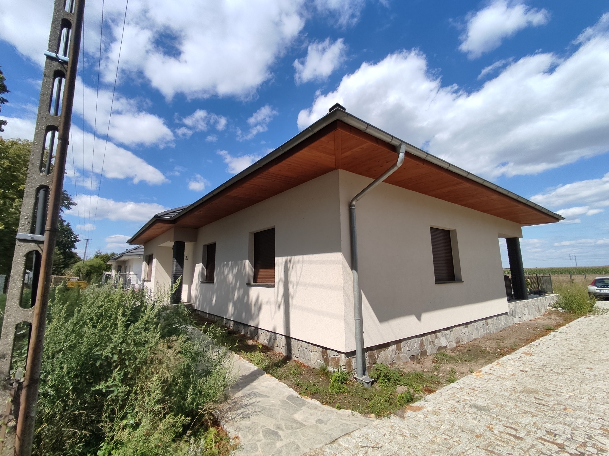 Nowoczesny, parterowy dom jednorodzinny z kremową elewacją, drewnianą podbitką dachu i kamienną podmurówką, widoczny z perspektywy chodnika z kostki brukowej w słoneczny dzień.