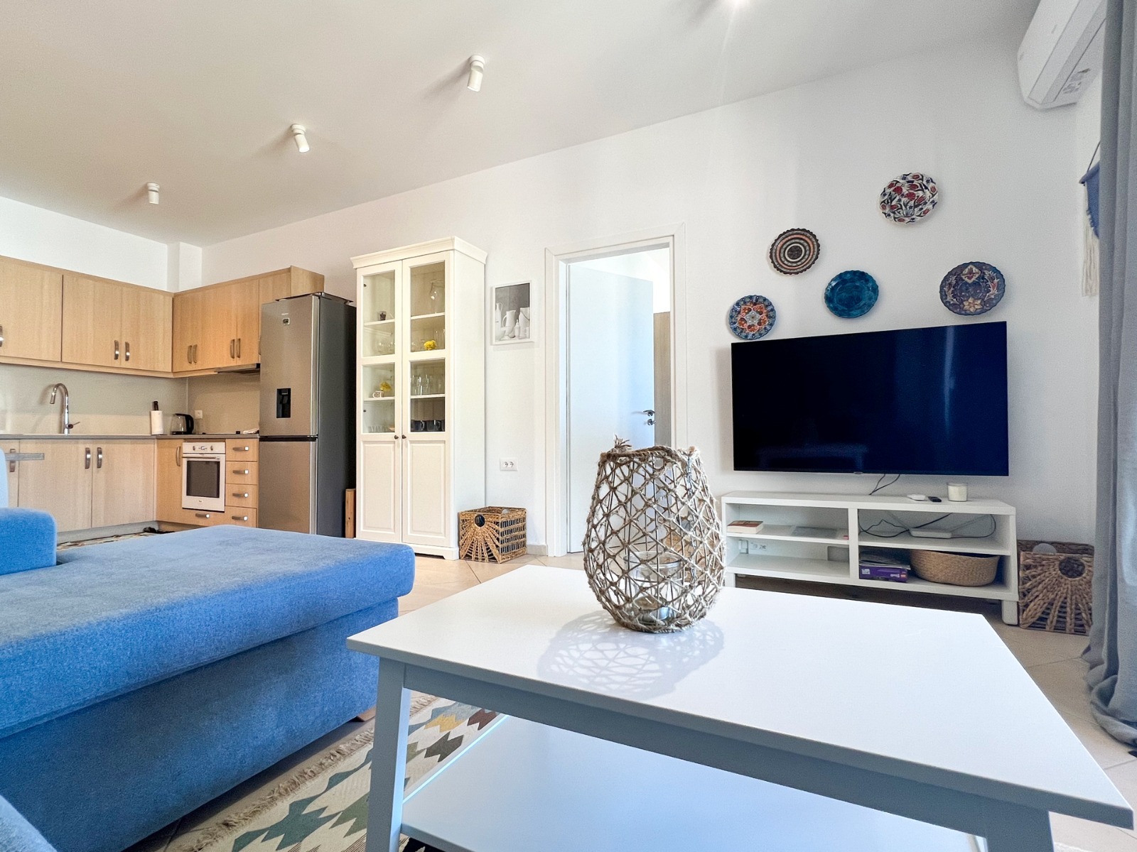 Jasne wnętrze salonu z aneksem kuchennym, niebieska sofa, biały stolik kawowy z dekoracyjnym lampionem, telewizor na ścianie z dekoracyjnymi talerzami, widoczna zabudowa kuchenna i szafka...