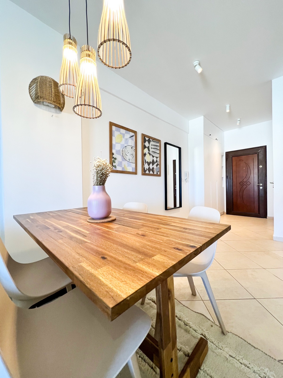 Jasne wnętrze jadalni z drewnianym stołem, białymi krzesłami, lampami wiszącymi z bambusa i dekoracjami ściennymi, widoczne płytki podłogowe i drzwi wejściowe.