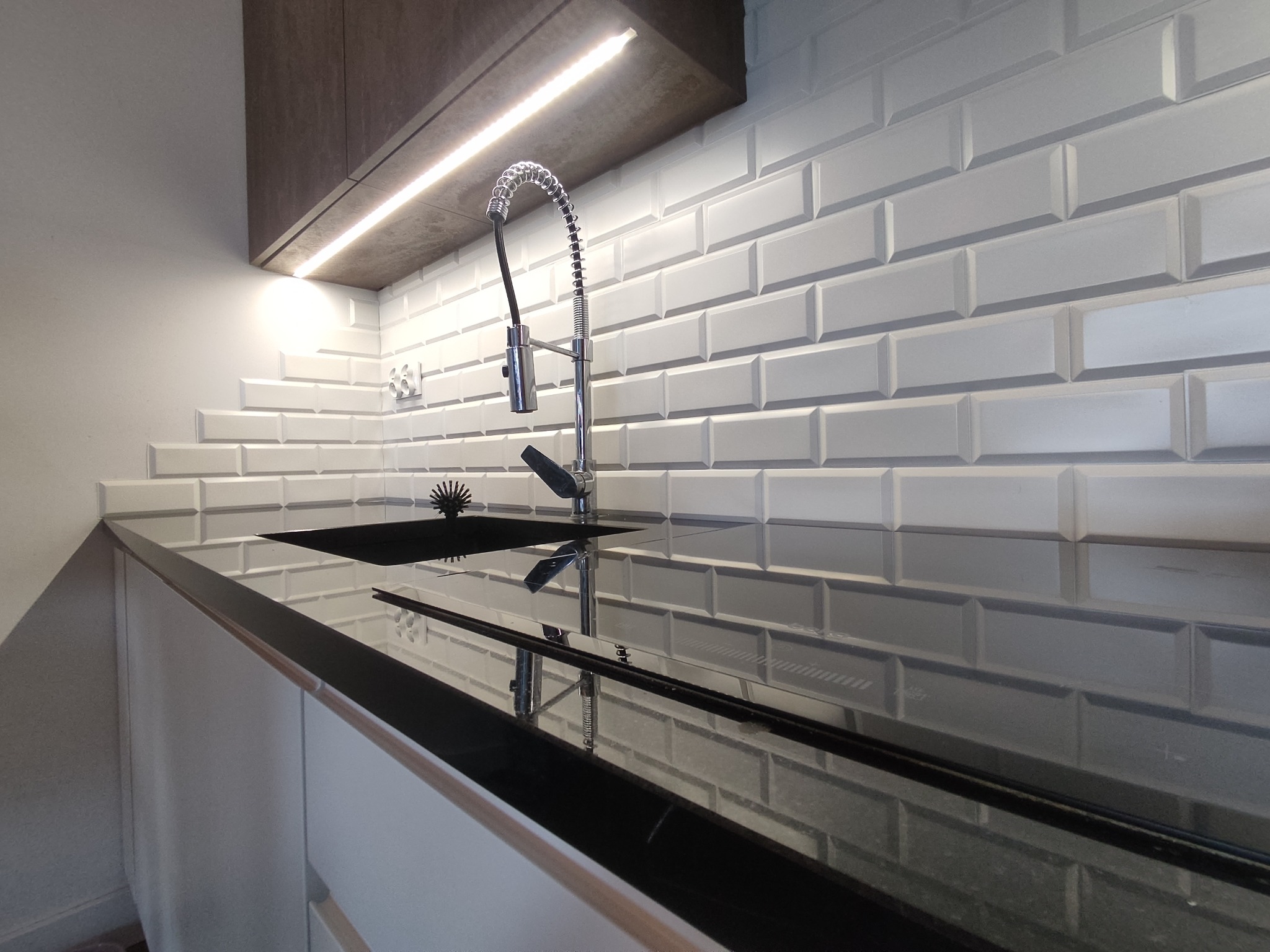 Białe, fazowane płytki ułożone w cegiełkę na ścianie nad czarnym, błyszczącym blatem kuchennym z wbudowanym zlewem i nowoczesną baterią ze sprężyną. Oświetlenie LED pod szafką górną.