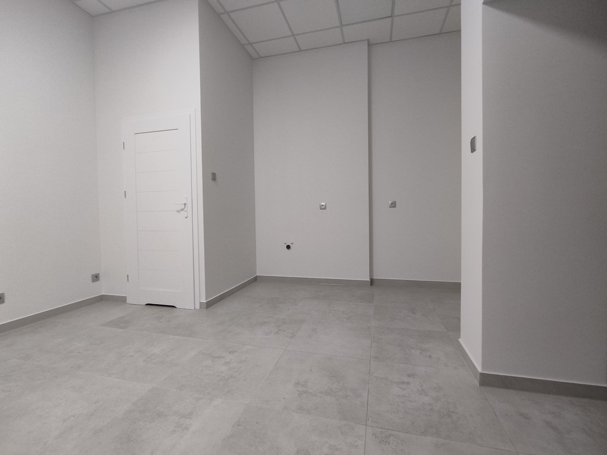 Puste, odnowione pomieszczenie z szarymi płytkami podłogowymi, białą ścianą i drzwiami w stylu minimalistycznym, widoczne gniazdka elektryczne.