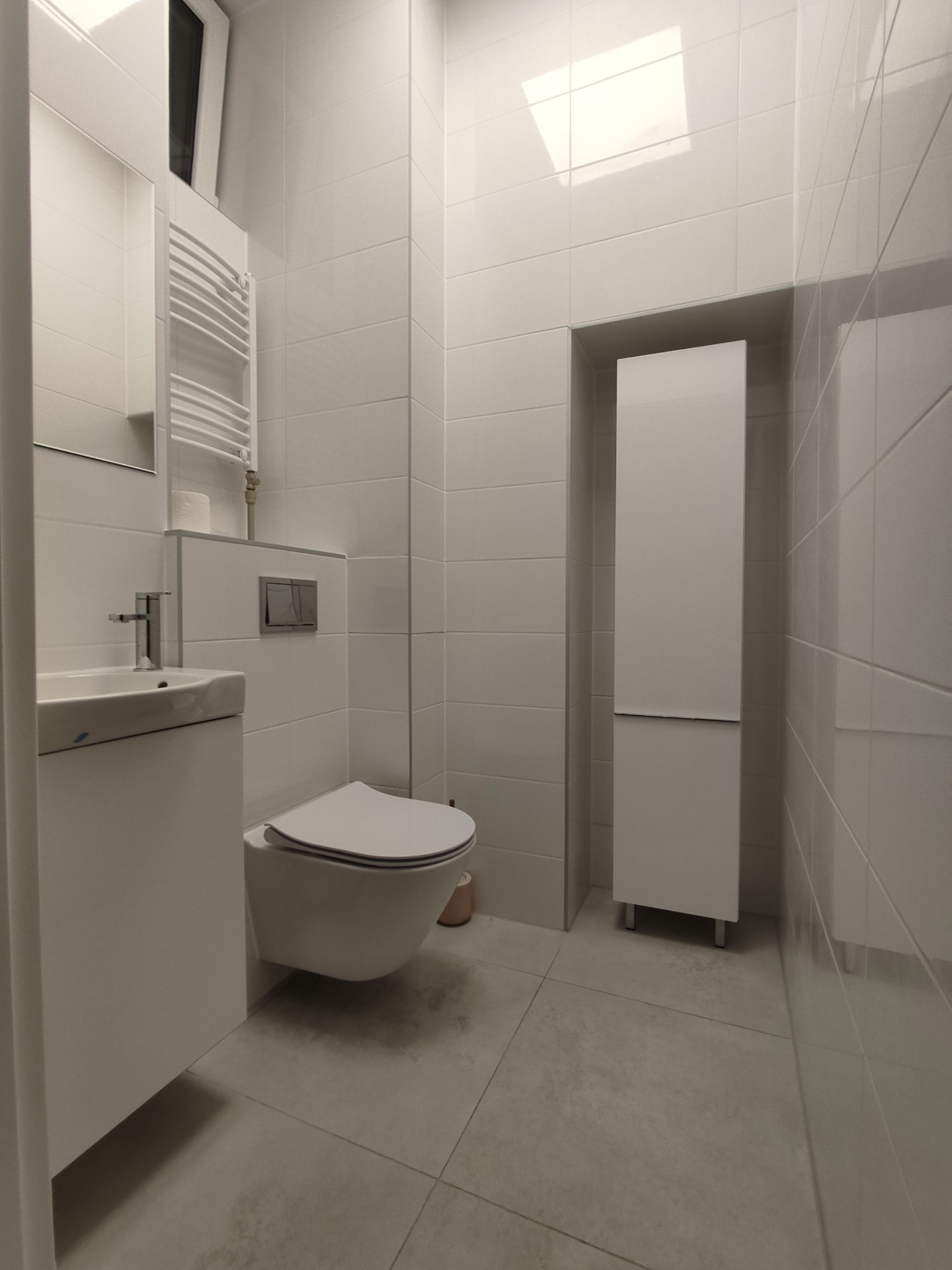 Nowoczesna, minimalistyczna łazienka z białymi płytkami, podwieszaną toaletą, umywalką z szafką i grzejnikiem drabinkowym. Widoczna wysoka szafka w zabudowie i szara podłoga.