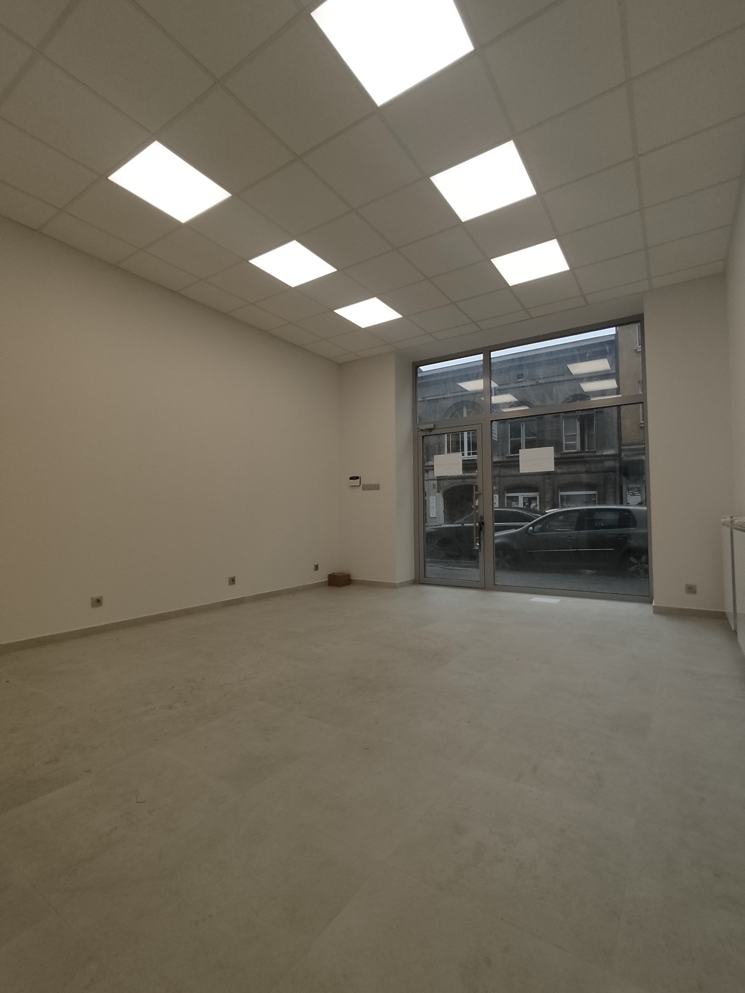 Puste, jasne pomieszczenie z białymi ścianami, płytkami podłogowymi w odcieniach szarości, podwieszanym sufitem z kwadratowymi lampami i dużymi szklanymi drzwiami wejściowymi z widokiem na ulicę.