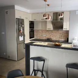 Jasna kuchnia z zabudową, wyspą z hokerami i miedzianymi lampami wiszącymi, widok na lodówkę z magnesami.