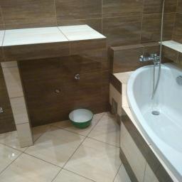 Gryfbud - Wykończona łazienka z brązowymi i beżowymi płytkami, biała wanna z chromowaną armaturą, umywalka w zabudowie z blatem i zielona miska na podłodze.