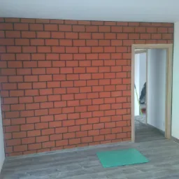 Wykończona ściana z efektem cegły w odcieniach czerwieni i szarości w mieszkaniu w Miechowie, widoczne panele podłogowe i ościeżnica drzwi.