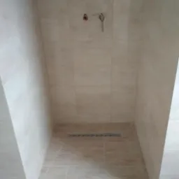 Wykończona kabina prysznicowa z beżowymi płytkami, odpływ liniowy w podłodze, widoczne przyłącza wodne na ścianie, dekoracyjny pas na górze ściany.