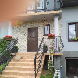 Wejście do domu z brązowymi drzwiami i schodami wyłożonymi płytkami, balustrada z metalu, donice z kwiatami, elewacja z kamienia dekoracyjnego, w tle otwarty garaż.