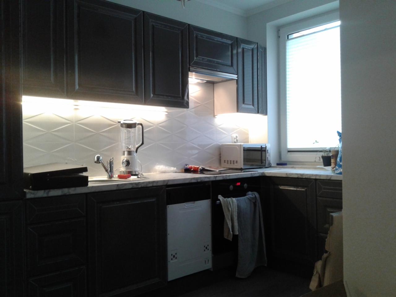 Ciemne szafki kuchenne z białymi romboidalnymi płytkami nad blatem, oświetlenie LED podszafkowe, mikser, zlew i kuchenka mikrofalowa widoczne na blacie, okno z żaluzjami w tle.