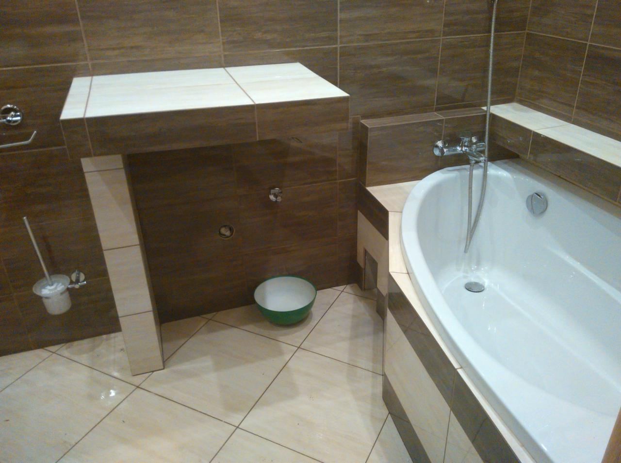 Wykończona łazienka z brązowymi i beżowymi płytkami, biała wanna z chromowaną armaturą, umywalka w zabudowie z blatem i zielona miska na podłodze.