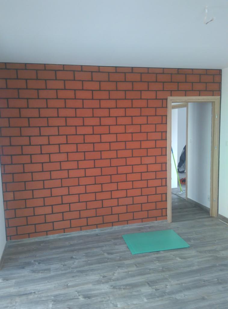 Wykończona ściana z efektem cegły w odcieniach czerwieni i szarości w mieszkaniu w Miechowie, widoczne panele podłogowe i ościeżnica drzwi.