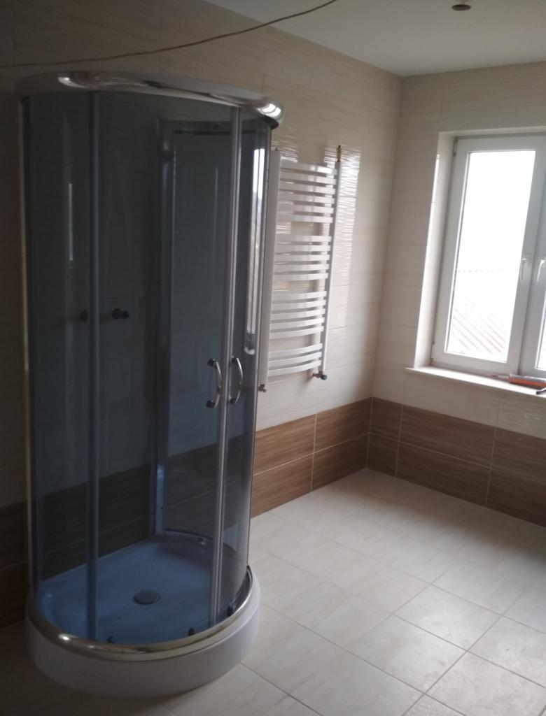 Zaokrąglona kabina prysznicowa z szarymi szybami i chromowanymi okuciami, grzejnik drabinkowy na ścianie z beżowo-brązowymi płytkami, białe płytki podłogowe, okno z białymi ramami w tle.