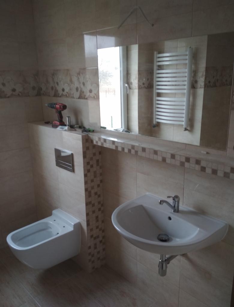 Montaż podwieszanej toalety i umywalki w łazience wykończonej beżowymi płytkami, widoczny grzejnik drabinkowy i narzędzia na półce.