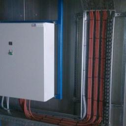 el-transfer Usługi elektryczno-budowlane Adam Duda - Szara skrzynka elektryczna z przyciskami, zamontowana na niebieskiej konstrukcji wsporczej, obok pionowe korytka z kablami w pomarańczowej izolacji, przymocowane do szarej ściany.