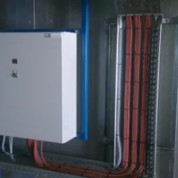 Szara skrzynka elektryczna z przyciskami, zamontowana na niebieskiej konstrukcji wsporczej, obok pionowe korytka z kablami w pomarańczowej izolacji, przymocowane do szarej ściany.