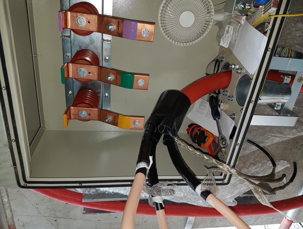 Wnętrze skrzynki elektrycznej z podłączonymi przewodami wysokiego napięcia, widoczne miedziane szyny zbiorcze oznaczone kolorami, wentylator i narzędzia.