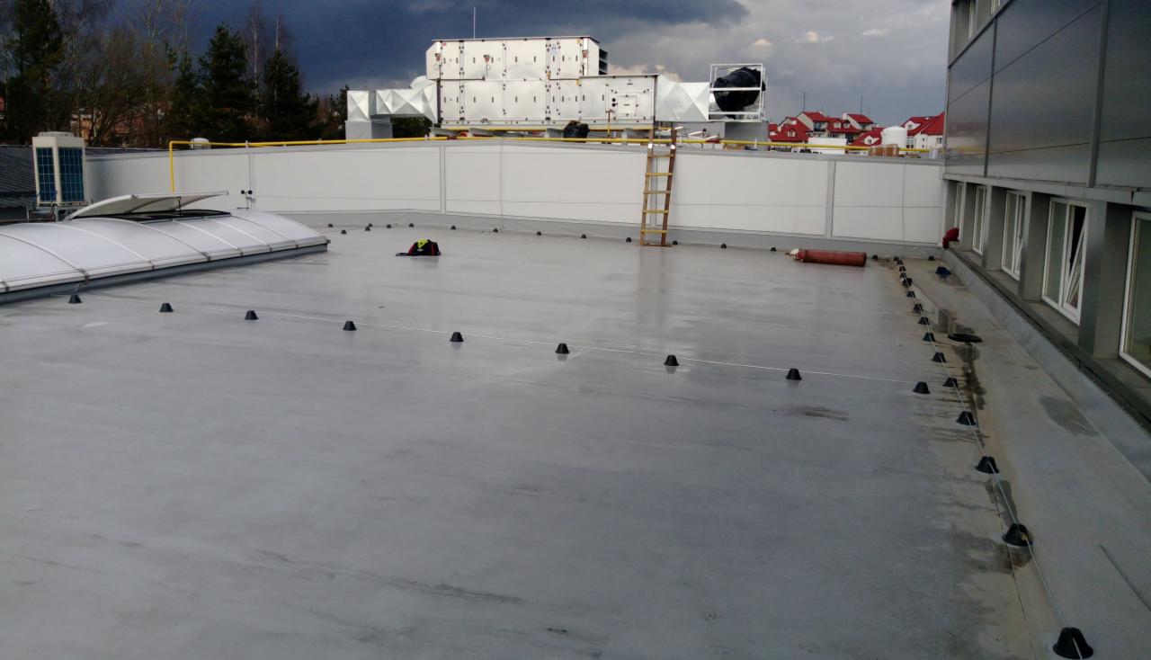 Płaski dach przemysłowy z instalacją odgromową w trakcie montażu, widoczne czarne uchwyty i linka odgromowa, drabina oparta o ścianę, w tle elementy wentylacji i pochmurne niebo.