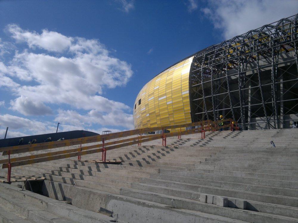 Stadion w budowie, widoczne trybuny z betonowych stopni, fragment elewacji w kolorze złota oraz konstrukcja stalowa dachu, pracownicy w kaskach na górze trybun.