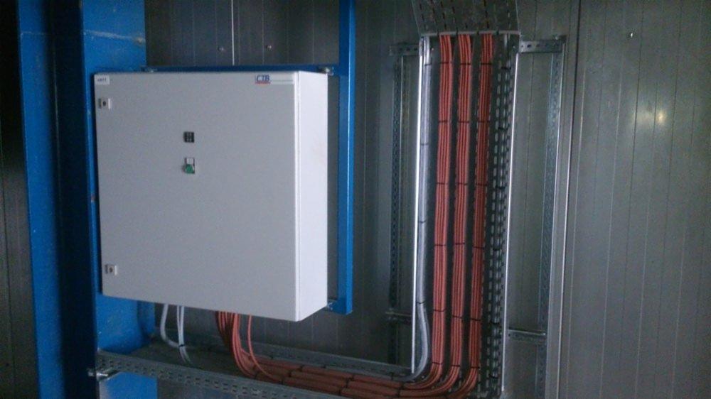 Szara skrzynka elektryczna z przyciskami, zamontowana na niebieskiej konstrukcji wsporczej, obok pionowe korytka z kablami w pomarańczowej izolacji, przymocowane do szarej ściany.