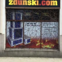 Fasada budynku z szyldem 'zdunski.com' i witryną z grafiką przedstawiającą przekrój pieca kaflowego, zabezpieczona metalową kratą o geometrycznym wzorze.