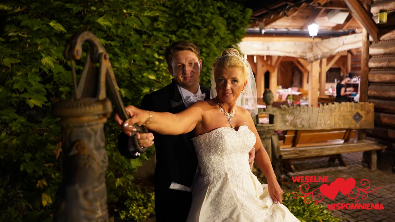 Uśmiechnięta para młoda pozuje przy starej studni na tle drewnianej konstrukcji i zieleni.